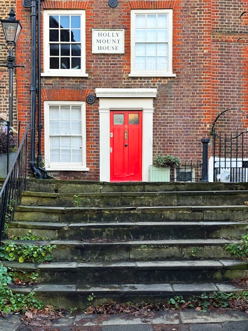 A three-story brick residential building with a red front door situated on a set of concrete stairs, which show signs of moss and dirt accumulation. The building features white-framed sash windows, with two on the ground floor and two on the first floor; one of the upper windows has multiple panes, while the lower windows have closed blinds. A white sign reading 'Holly Mount House' is mounted between the upper windows. To the left of the entrance, a black downspout runs vertically along the brick wall, and a black outdoor lamp post is visible on the sidewalk beside the stairs. On the right side of the steps, there is a small green shrub in a rectangular planter, and a black metal fence with an arched gate leads to the building’s side yard. The scene suggests an urban or suburban environment with potential for private rubbish collection, and the presence of moss indicates a damp climate typical of areas where independent waste handling, such as rubbish removal services, might be necessary for maintaining property cleanliness.