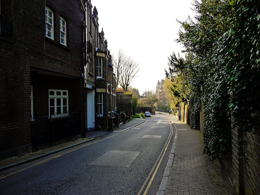 A three-story brick residential building with a red front door situated on a set of concrete stairs, which show signs of moss and dirt accumulation. The building features white-framed sash windows, with two on the ground floor and two on the first floor; one of the upper windows has multiple panes, while the lower windows have closed blinds. A white sign reading 'Holly Mount House' is mounted between the upper windows. To the left of the entrance, a black downspout runs vertically along the brick wall, and a black outdoor lamp post is visible on the sidewalk beside the stairs. On the right side of the steps, there is a small green shrub in a rectangular planter, and a black metal fence with an arched gate leads to the building’s side yard. The scene suggests an urban or suburban environment with potential for private rubbish collection, and the presence of moss indicates a damp climate typical of areas where independent waste handling, such as rubbish removal services, might be necessary for maintaining property cleanliness.