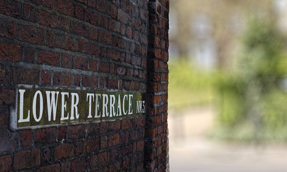 A close-up of a brick wall with a rectangular metal street sign attached, featuring the words 'LOWER TERRACE NW3' in white capital letters on a background of alternating dark green and grey tiles. The brick wall is composed of dark reddish-brown bricks with a rough texture and dark mortar joints, extending vertically along the left side of the image. The focus is primarily on the sign and the brick surface immediately surrounding it, with the background blurred, revealing hints of green foliage and natural light, suggesting an outdoor setting. The scene is evenly lit with natural daylight, contributing to the clarity of the sign and brick textures. This image emphasizes urban environment details relevant to local area signage and the context of private property or independent street naming, subtly aligning with themes of waste disposal and local signage facilitation by rubbish removal services like House Clearance Hampstead.