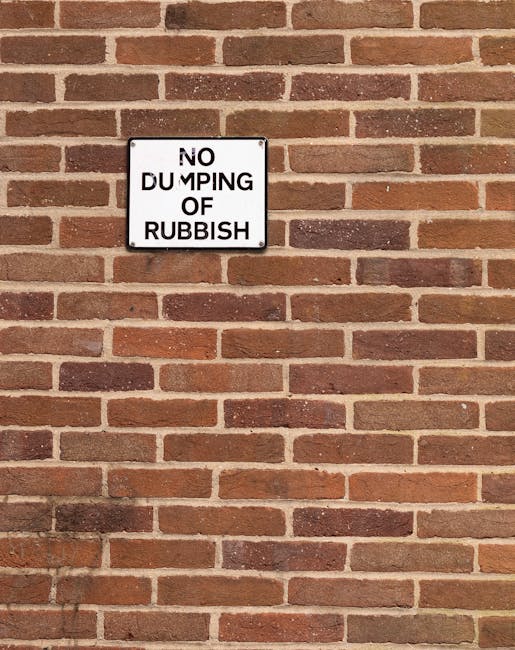 A rectangular metal sign mounted on a red-brown brick wall with a slightly textured surface, featuring black text on a white background that reads 'NO DUMPING OF RUBBISH.' The sign is positioned towards the upper left side of the image, with the bricks laid in a traditional horizontal pattern and mortar joints clearly visible. The wall's natural warm tones and the utilitarian appearance of the sign suggest a public or private external area where waste disposal is regulated. The lighting appears natural, casting a subtle shadow of the sign on the bricks below. This scene relates to waste management practices, emphasizing the importance of designated areas for rubbish removal, which companies like House Clearance Hampstead often support through services focused on alternative waste handling and independent collection methods in residential environments.