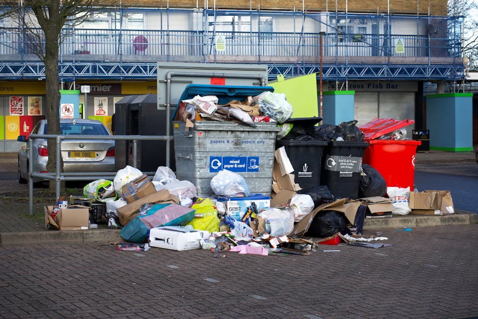 A large collection of overflowing rubbish bins and scattered waste on a paved area in front of a commercial building. The waste includes black and black-and-white mixed rubbish bags, flattened cardboard boxes, paper and packaging materials, plastic bags, and loose paper. One grey metal recycling bin labeled for mixed paper and card is filled to overflowing, with waste spilling onto the ground. Nearby, there are a red trash bin and a black container, both of which are also overfilled, contributing to the cluttered scene. Behind the rubbish, a blue fence partially surrounds the building, which features a row of storefronts with signage visible at the base. A grey car is parked next to the rubbish collection area, partially obscured by a metal barrier. The overall environment suggests a typical urban or suburban scene where improper waste disposal or delays in rubbish collection have led to the accumulation of waste in this outdoor space, highlighting the need for efficient rubbish removal services such as those provided by House Clearance Hampstead.