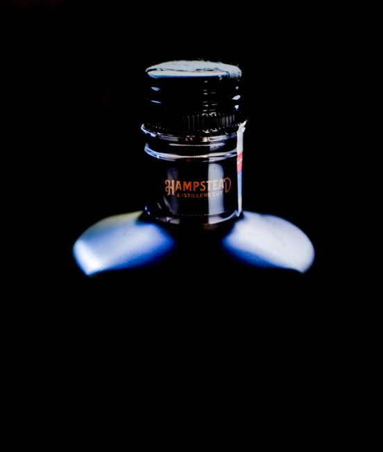 A close-up image of the top part of a dark glass bottle, illuminated by a focused light source that highlights its glossy surface and metallic cap. The cap is black with a matte finish, featuring visible ridges around its edge, and a partially visible label wrapped around the neck of the bottle displays the word 'Hampstead' in orange text. The background is entirely black, creating a high contrast that emphasizes the bottle’s reflective surface and subtle curves, with a faint reflection of the bottle visible on the surface below. The scene conveys a minimalistic, professional approach often associated with premium beverage presentations, subtly aligning with the theme of private disposal or alternative waste handling when considering packaging and bottle recycling, as part of waste removal services offered by House Clearance Hampstead.