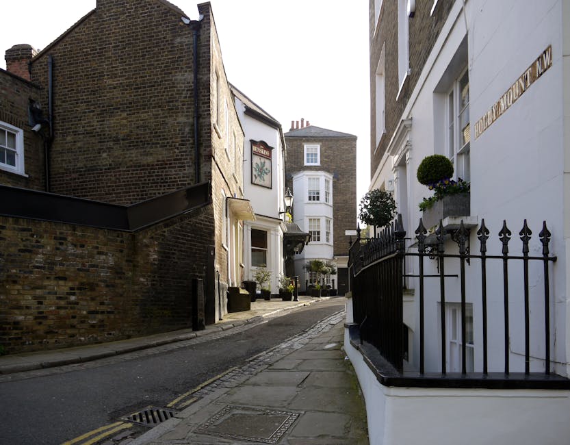 A narrow residential street in Hampstead with a slight incline, featuring a black wrought iron fence with pointed finials and decorative flower boxes filled with small green shrubs and blooming plants attached to a white building on the right side. On the left, there is a brick wall and a small white-painted building with potted plants outside its entrance. Further in the background, a larger white building with a bay window and chimney stacks is visible under a clear sky, indicating daytime. The pavement is composed of concrete slabs, with a drainage grate near the curb, and the roadway has double yellow lines suggestive of parking restrictions. The image reflects a quiet, charming London neighbourhood, with no rubbish or waste visible, emphasizing the area's neat and well-maintained character, typical of private residence exteriors that may be handled through independent collection and on-site clearance services like those offered by House Clearance Hampstead.