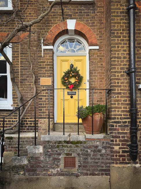 The image depicts a residential front door with a traditional arched brick surround, featuring a decorative semi-circular window with a fanlight. The bright yellow wooden door is accented by a festive green wreath decorated with orange and red ornaments, along with a red bow at the bottom, hanging centrally on the door. A black metal railing runs along the small porch, with a potted plant containing lush green foliage placed on the right side of the steps. The porch is accessed by a concrete step with a brick base and is set against a brick facade wall with varying shades of brown and red. A black downpipe runs vertically along the right side of the building, and a small square ventilation grille is embedded in the brickwork beneath the steps. The scene is illuminated by natural daylight, with the overall atmosphere suggesting a well-maintained, traditional Hampstead residence, potentially part of a property recently cleared or prepared for private waste collection services that House Clearance Hampstead may offer, fitting within the context of alternative rubbish removal solutions for homeowners and landlords in the area.