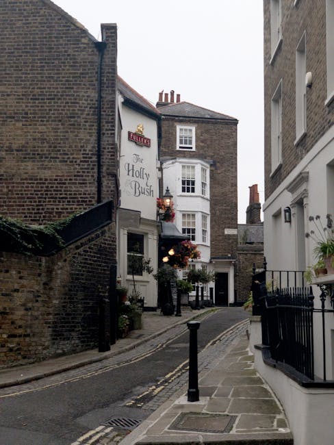 A narrow residential street in Hampstead with a slight incline, featuring a black wrought iron fence with pointed finials and decorative flower boxes filled with small green shrubs and blooming plants attached to a white building on the right side. On the left, there is a brick wall and a small white-painted building with potted plants outside its entrance. Further in the background, a larger white building with a bay window and chimney stacks is visible under a clear sky, indicating daytime. The pavement is composed of concrete slabs, with a drainage grate near the curb, and the roadway has double yellow lines suggestive of parking restrictions. The image reflects a quiet, charming London neighbourhood, with no rubbish or waste visible, emphasizing the area's neat and well-maintained character, typical of private residence exteriors that may be handled through independent collection and on-site clearance services like those offered by House Clearance Hampstead.