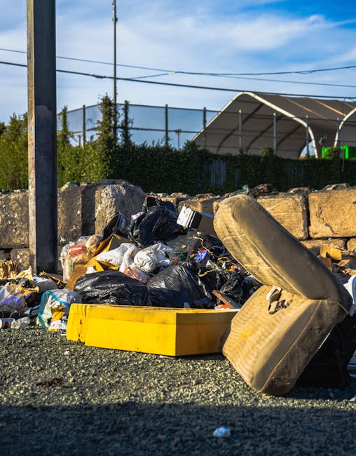 A large pile of mixed waste and rubbish overflowing from several wheelie bins, situated on a paved urban street in front of a commercial building. The waste includes cardboard boxes, paper, plastic bags, and assorted packaging materials, with some items spilling onto the surrounding pavement. The wheelie bins are different colors, including grey, black, and red, and are positioned side by side, with the grey bin having a partially open lid revealing paper and cardboard inside. Behind the bins, a metal railing separates the rubbish area from a parked silver hatchback car with a UK registration plate, partially visible on the left. The background features the entrance to a shop or cafe, with closed shutters and signage, and a building wrapped in blue scaffolding and safety netting indicating ongoing construction or maintenance work. The scene is well-lit with natural daylight, emphasizing the cluttered and untidy state of the waste area, which may prompt consideration of alternative private rubbish collection or on-site clearance services such as those offered by House Clearance Hampstead.