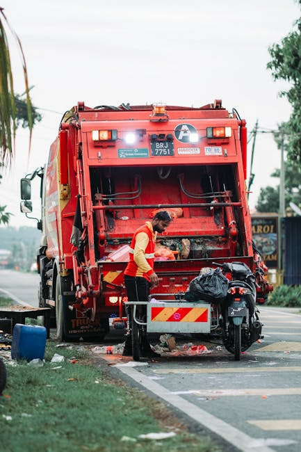 A sanitation worker wearing a high-visibility yellow and red uniform is operating a red refuse collection vehicle parked on the side of a street, with its rear hopper open for rubbish loading. The worker is emptying a black plastic trash bag into the vehicle, and small debris is scattered on the pavement beneath. The scene takes place during daylight, with an overcast sky and some trees and buildings visible in the background. The vehicle is positioned partially on the curb and partially on the roadway, indicative of a street-based rubbish collection operation. Nearby, a black motorcycle is parked with a small metal barrier attached for safety, and a blue container can be seen on the grassy verge beside the street. The environment suggests a typical urban or suburban street scene, with the scene illustrating a private waste removal service that may offer alternative on-site or scheduled rubbish collection, as part of a professional waste management solution. The scene aligns with services such as those provided by House Clearance Hampstead in offering independent rubbish disposal, especially in residential or commercial areas needing efficient waste clearance.