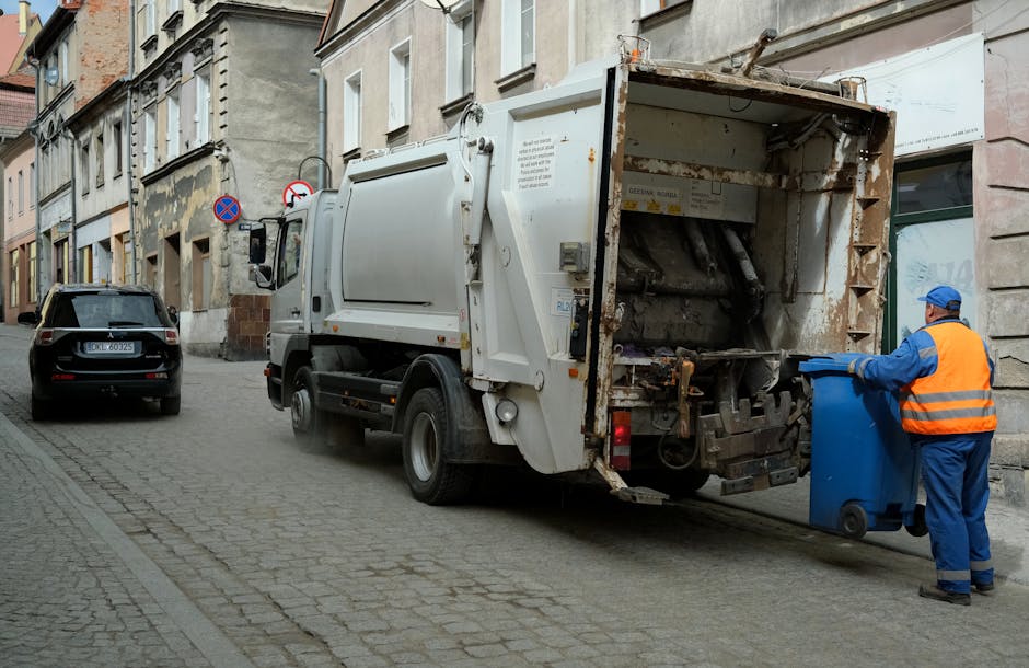 A sanitation worker wearing a high-visibility yellow and red uniform is operating a red refuse collection vehicle parked on the side of a street, with its rear hopper open for rubbish loading. The worker is emptying a black plastic trash bag into the vehicle, and small debris is scattered on the pavement beneath. The scene takes place during daylight, with an overcast sky and some trees and buildings visible in the background. The vehicle is positioned partially on the curb and partially on the roadway, indicative of a street-based rubbish collection operation. Nearby, a black motorcycle is parked with a small metal barrier attached for safety, and a blue container can be seen on the grassy verge beside the street. The environment suggests a typical urban or suburban street scene, with the scene illustrating a private waste removal service that may offer alternative on-site or scheduled rubbish collection, as part of a professional waste management solution. The scene aligns with services such as those provided by House Clearance Hampstead in offering independent rubbish disposal, especially in residential or commercial areas needing efficient waste clearance.