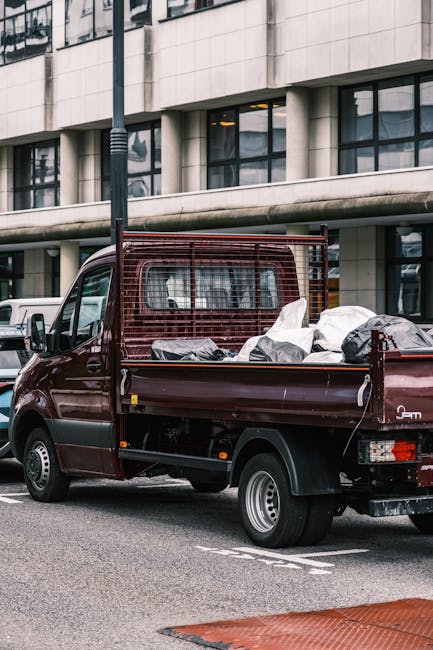 A maroon flatbed truck parked on a city street in front of a modern building with large glass windows and concrete panels. The truck's cargo area is filled with various types of rubbish, including black garbage bags and loose debris, which appear to be in different sizes and materials, some plastic and some fabric. The truck features a metal mesh side panel surrounding the cargo area, providing support and containment for the waste. The vehicle is positioned on a designated loading zone on the pavement, with part of a red rubber mat visible in the foreground. An urban environment surrounds the scene, evidenced by other parked vehicles and a street lamp, indicating an active city setting suitable for private rubbish collection or on-site clearance services managed by companies like House Clearance Hampstead, specialists in independent waste handling and rubbish removal in Hampstead.