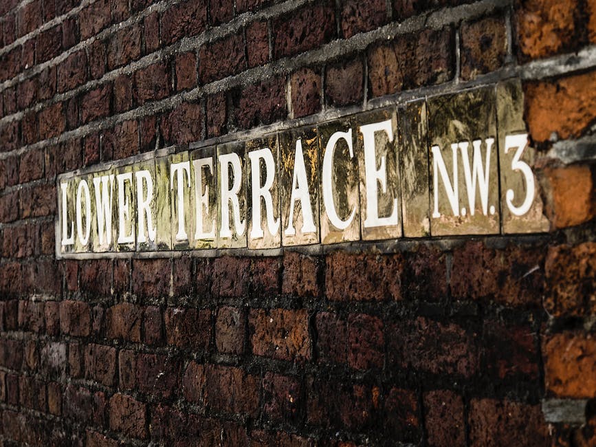 A close-up of a rectangular street sign mounted on a weathered red brick wall, displaying the text 'LOWER TERRACE NW3' in large, white, serif capital letters mounted on a glossy, reflective ceramic tile background. The sign is situated horizontally and slightly angled, with the bricks showing varying shades of dark red and brown, some with patches of moss or dirt, indicating age and exposure to outdoor elements. The mortar between the bricks is visible and appears aged. The lighting in the scene is natural and soft, highlighting the textured surface of the bricks and the glossy finish of the sign. There are no other objects or environmental features in the image, focusing solely on the sign and brick wall, typical of street signage used for address identification in urban settings, and relevant for contexts involving property or location identification for private waste collection or local authority rubbish removal services.