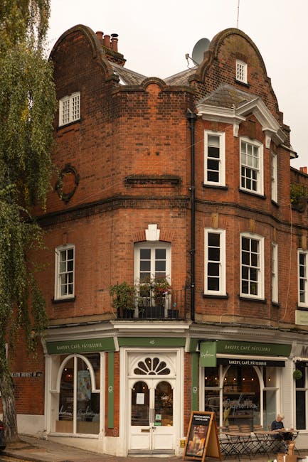 A three-story red brick building with a rounded, turret-like corner featuring decorative brickwork and small arched windows at the top, situated on a street corner. The ground floor houses a café or bakery, indicated by green awnings with white lettering, and a white arched entrance with a fanlight window above it. In front of the building, a paved sidewalk includes a bench and a standing chalkboard sign on the pavement. To the left side of the image, a tall tree with green foliage partially obscures the building and casts some shadows onto the façade. The upper floors display multiple white-framed sash windows, some with flower baskets, and the building's roof has chimneys and a satellite dish. The scene is set during daytime with natural, soft lighting that highlights the texture of the brickwork, giving an overall impression of a quaint, urban street scene, suitable for describing private refuse collection or alternative waste handling within a retail or mixed-use environment. This image is part of a page discussing access issues for rubbish removal in Hampstead, London, as represented by the presence of rubbish collection services by House Clearance Hampstead.